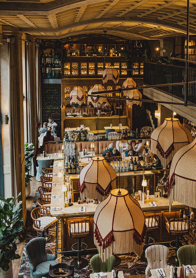 Stimmungsvolle Gesamtaufnahme des GIGI Restaurants in München mit imposanten Fransenlampen, offener Bar, eleganten Sitzbereichen und stilvoller Lichtinszenierung – italienisches Flair auf zwei Ebenen.