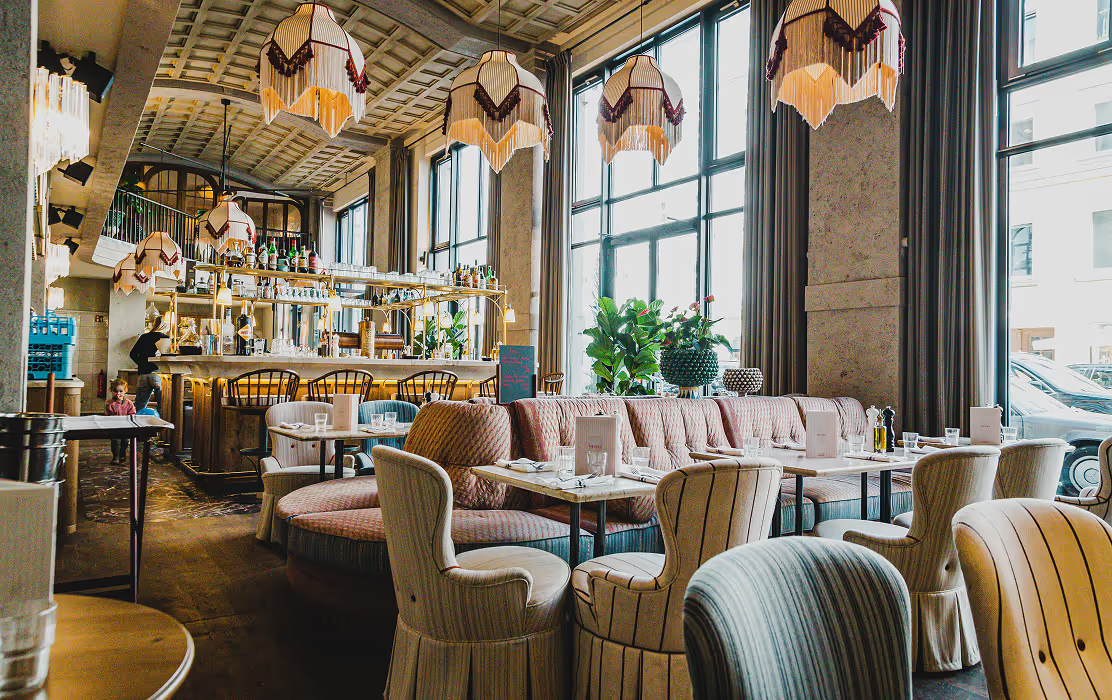 Heller, stilvoll eingerichteter Gastraum der GIGI Trattoria in München mit großen Fensterfronten, rosafarbenen Samtsofas, gemusterten Sesseln und Retro-Lampen – italienisches Flair in modernem Ambiente.