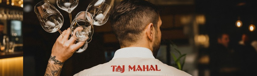 Servicekraft im Taj Mahal Restaurant mit aufgesticktem Logo auf der Rückenpartie der Uniform – stilvoll balanciert sie mehrere Weingläser mit der Hand.